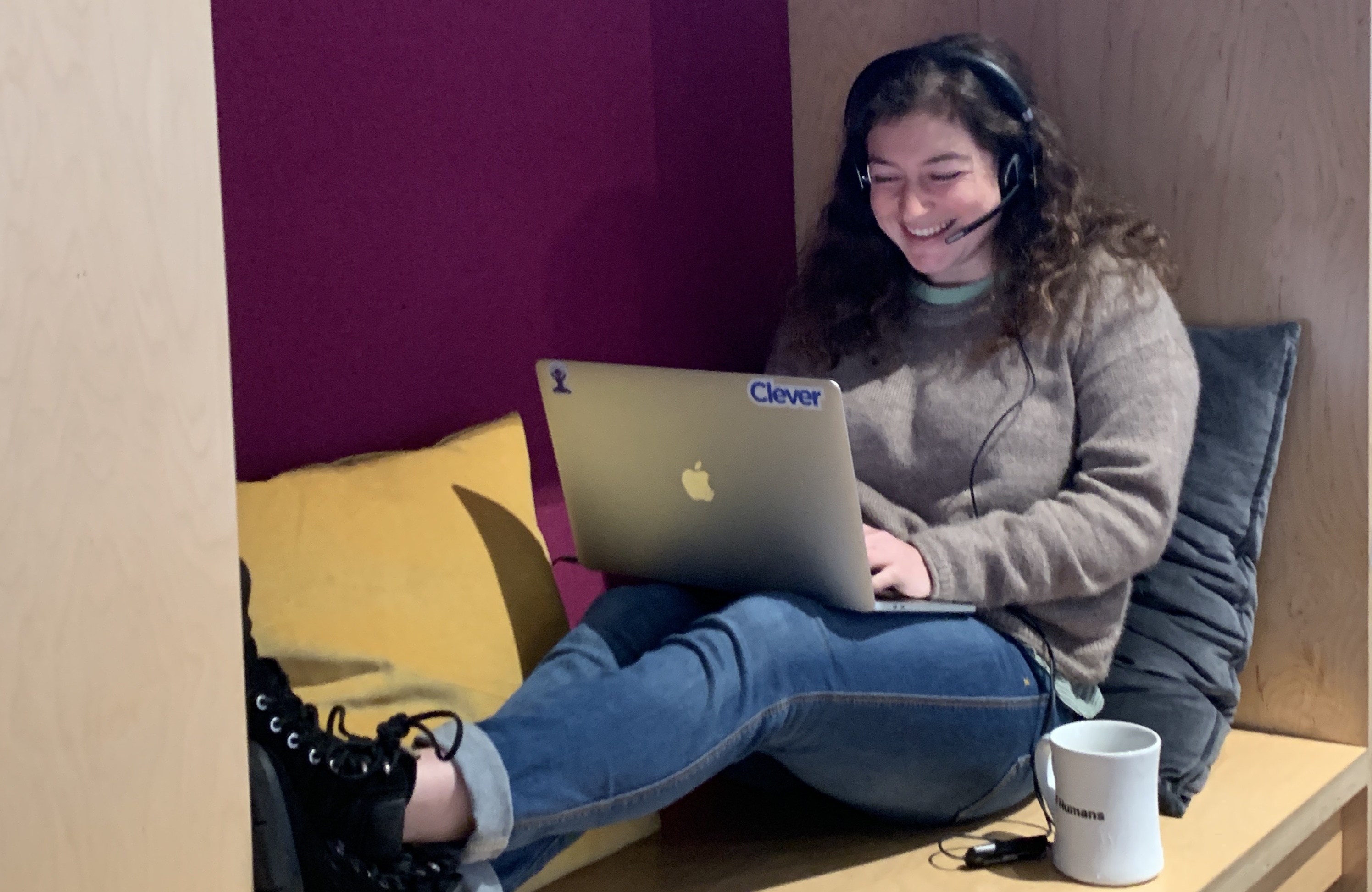 A photo of Sarah Markowitz, lounging in a wooden seating structure and working on her Clever laptop with a phone headset. 